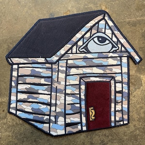 May include: A house-shaped rug featuring a camouflage design in blue, grey, and white. The roof is navy blue, and the door is burgundy. An eye symbol is above the door. The rug is designed to resemble a small cabin.