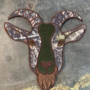 May include: A goat head patch with a camouflage pattern, brown horns, and a green center. The patch has a brown border and is likely for a jacket or bag.
