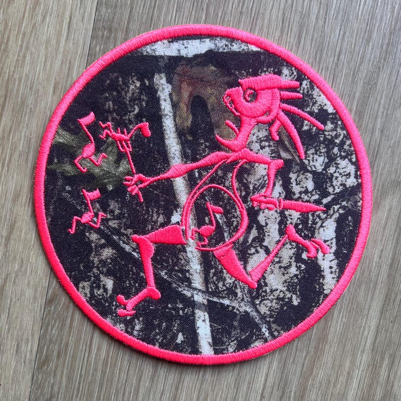 May include: A circular patch with a pink border and a camouflage pattern. A pink figure with musical notes is in the centre. The figure is holding a staff with musical notes and has a musical note symbol on its body.