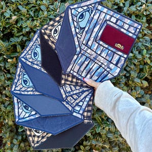 K&ouml;nnte beinhalten: Eine Clutch mit geometrischem Muster in Blau-, Schwarz- und Beiget&ouml;nen, mit Augen- und Schnurrbart-Designs. Die Clutch hat eine rechteckige Form mit mehreren Paneelen und einem burgunderfarbenen Akzent.