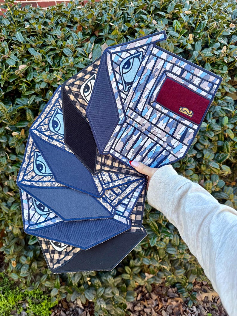 K&ouml;nnte beinhalten: Eine Clutch mit geometrischem Muster in Blau-, Schwarz- und Beiget&ouml;nen, mit Augen- und Schnurrbart-Designs. Die Clutch hat eine rechteckige Form mit mehreren Paneelen und einem burgunderfarbenen Akzent.