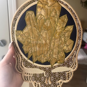 以下が含まれることがあります： クリスタル形成を特徴とする様式化されたスカルの刺繍パッチ。デザインには、金色の結晶のクラスターが付いた濃い青色の円が含まれています。スカルとボーダーは、幾何学模様のタン色です。