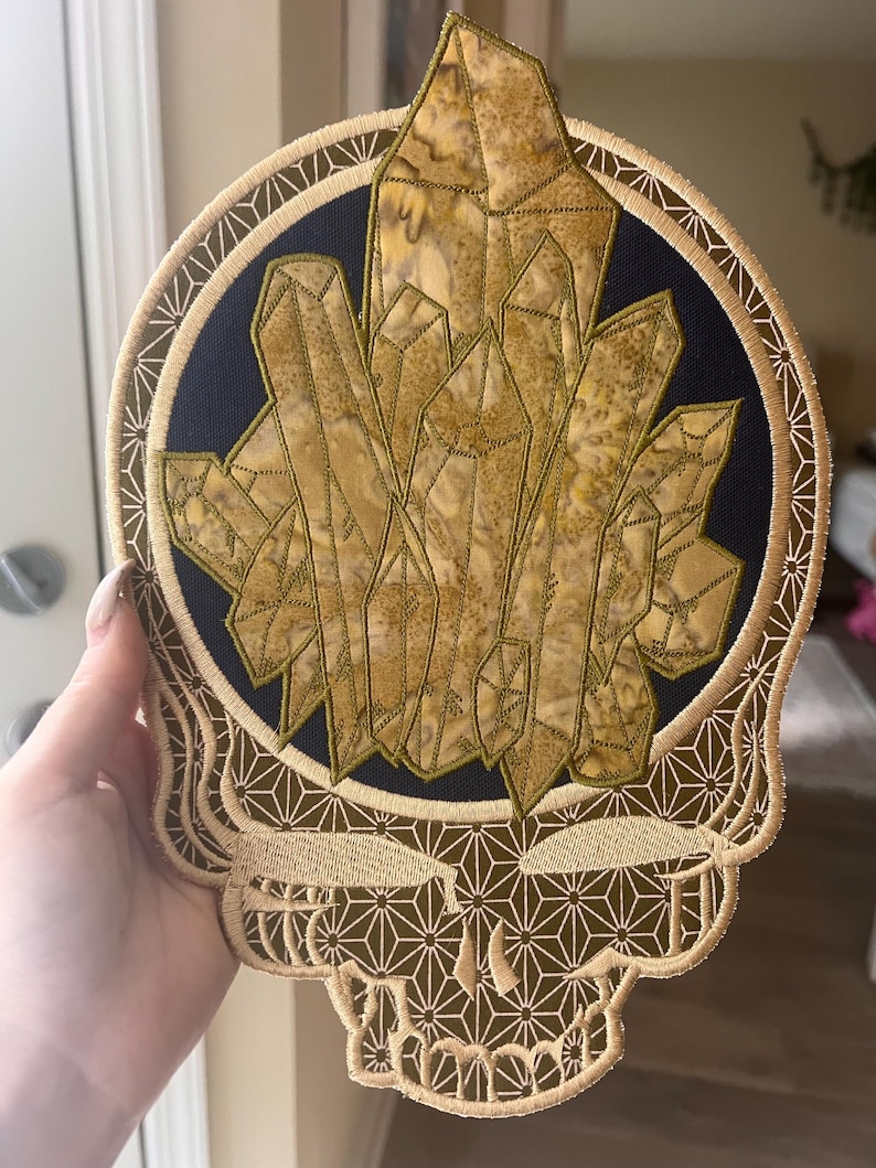 Pu&ograve; includere: Toppa ricamata con un teschio stilizzato con una formazione di cristalli. Il design include un cerchio blu scuro con un gruppo di cristalli dorati. Il teschio e il bordo sono di colore beige con motivi geometrici.