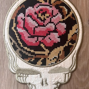 以下が含まれることがあります： 刺繍パッチ。バラのデザインのスタイリッシュな頭蓋骨が特徴です。バラはピンクと赤の色合いで、黒い背景に配置されています。頭蓋骨は白で、金と茶色のアクセントが付いています。ユニークな装飾アイテムです。