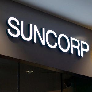 以下が含まれることがあります： 暗い灰色の壁に「SUNCORP」と書かれた白い文字が光っています。看板はガラス張りの店の入り口の上に設置されています。文字は3D効果があり、影を落としています。
