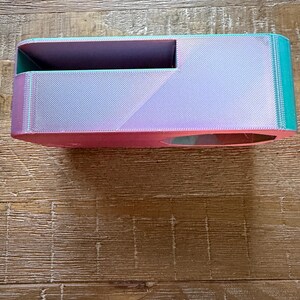 May include: A rectangular 3D printed storage container featuring a color-shifting gradient finish that transitions from vibrant pink to teal. The object has a wide, open top compartment and a smaller circular cutout on the side, showcasing a textured surface.
