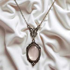 Puede incluir: Un collar de estilo antiguo con un colgante ovalado. El colgante está enmarcado en un metal color bronce con detalles ornamentados. La cadena y los elementos decorativos también son de bronce. El colgante parece contener una piedra o un inserto de color rosa pálido.