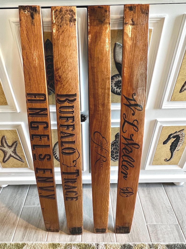 May include: Four reclaimed wood planks with dark lettering. The planks are engraved with the words "ANGELS ENVY", "BUFFALO TRACE", "E.H. Taylor", and "W.L. Weller". The wood has a natural, warm brown tone with visible grain.