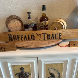 Puede incluir: Una exhibici&oacute;n con dos peque&ntilde;os barriles de madera, dos botellas de bourbon y un letrero de madera que dice "BUFFALO TRACE". El letrero es arqueado y tiene una silueta de b&uacute;falo. Las botellas son marrones y transparentes. La exhibici&oacute;n est&aacute; sobre un mueble blanco.