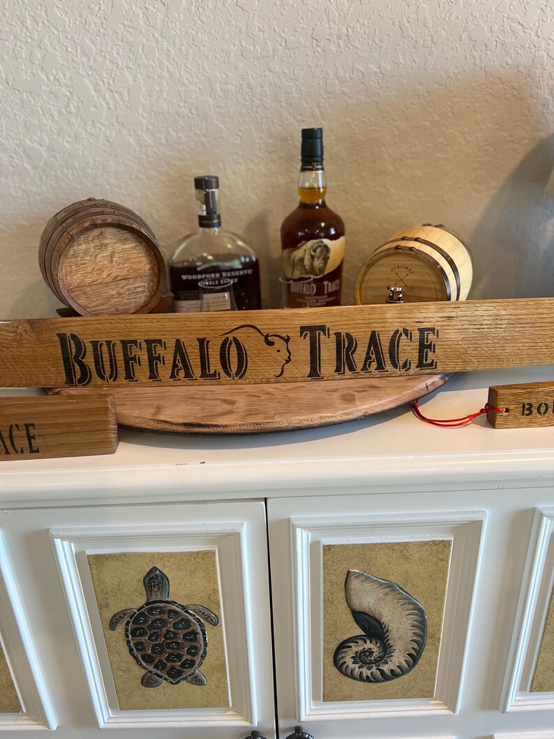 Puede incluir: Una exhibici&oacute;n con dos peque&ntilde;os barriles de madera, dos botellas de bourbon y un letrero de madera que dice "BUFFALO TRACE". El letrero es arqueado y tiene una silueta de b&uacute;falo. Las botellas son marrones y transparentes. La exhibici&oacute;n est&aacute; sobre un mueble blanco.