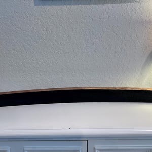 May include: A long, curved wooden shelf with a dark, black center and brown edges. The shelf is made from a barrel stave and is displayed on a white surface. The shelf is approximately 40 inches long.