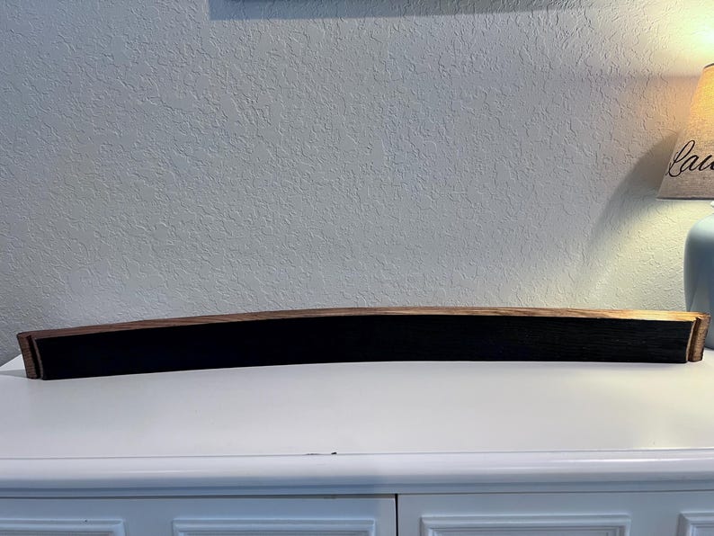 May include: A long, curved wooden shelf with a dark, black center and brown edges. The shelf is made from a barrel stave and is displayed on a white surface. The shelf is approximately 40 inches long.