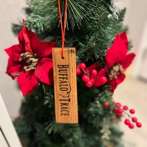May include: A wooden ornament with the text "BUFFALO TRACE" is suspended from a red string. The ornament is rectangular and hangs on a decorated Christmas tree with red poinsettias and berries.