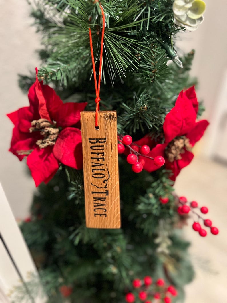 May include: A wooden ornament with the text "BUFFALO TRACE" is suspended from a red string. The ornament is rectangular and hangs on a decorated Christmas tree with red poinsettias and berries.