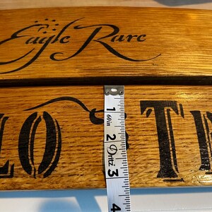 Op de afbeelding: Close-up van twee houten planken met de woorden "Eagle Rare" en "LOTR" in zwart geschilderd. De planken zijn ongeveer 5 cm breed. Het hout heeft een natuurlijke, warme bruine tint. Een meetlint is zichtbaar.