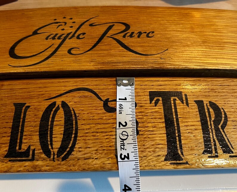 Op de afbeelding: Close-up van twee houten planken met de woorden "Eagle Rare" en "LOTR" in zwart geschilderd. De planken zijn ongeveer 5 cm breed. Het hout heeft een natuurlijke, warme bruine tint. Een meetlint is zichtbaar.