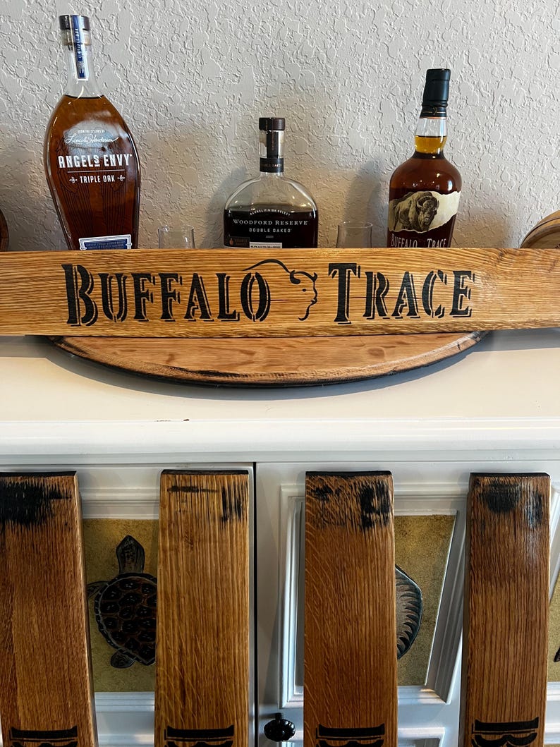 May include: A wooden sign with the words "BUFFALO TRACE" in black lettering. The sign is above a white cabinet with wooden panels. Three bottles of bourbon are visible, including Angel's Envy and Buffalo Trace.