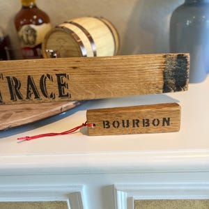 Puede incluir: Una duela de barril de bourbon de madera con la palabra "RACE" impresa. Una etiqueta de madera m&aacute;s peque&ntilde;a con la palabra "BOURBON" est&aacute; sujeta con una cuerda roja. Los art&iacute;culos son de madera marr&oacute;n claro.