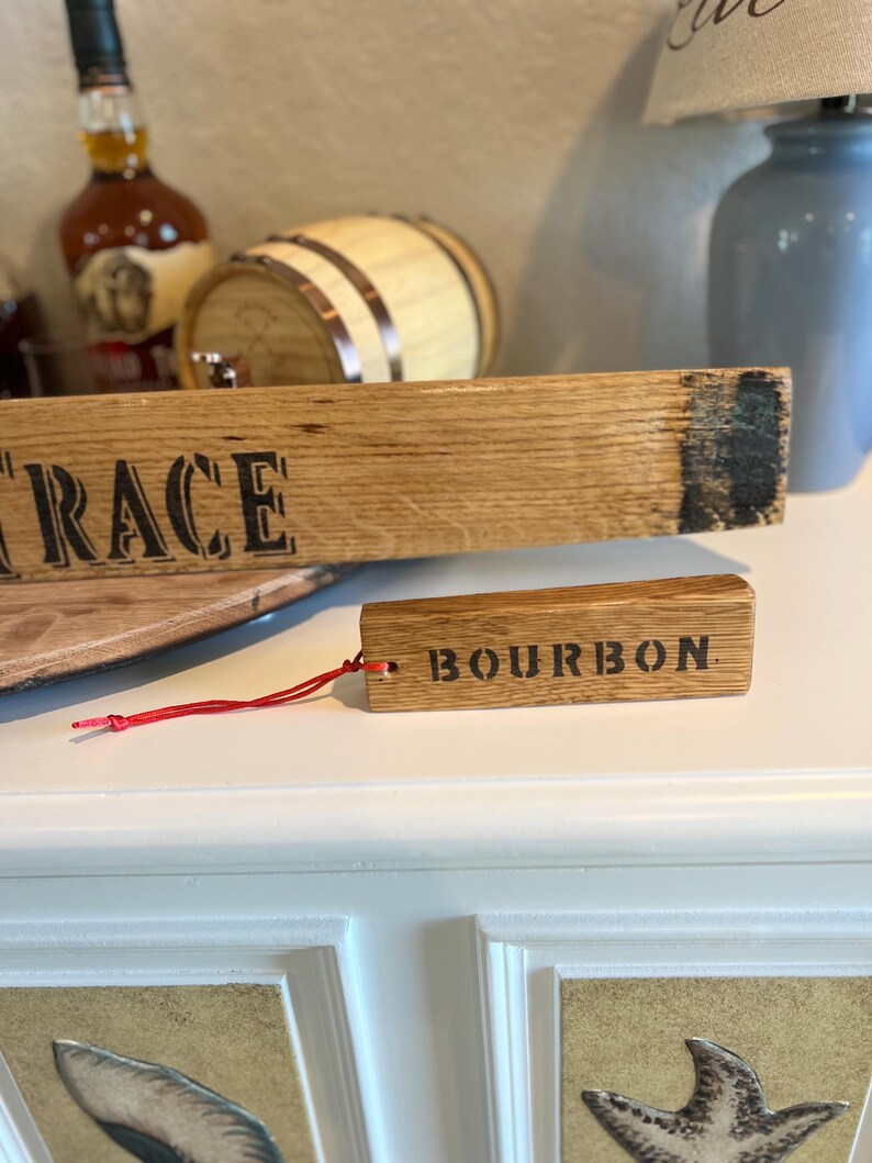 Puede incluir: Una duela de barril de bourbon de madera con la palabra "RACE" impresa. Una etiqueta de madera m&aacute;s peque&ntilde;a con la palabra "BOURBON" est&aacute; sujeta con una cuerda roja. Los art&iacute;culos son de madera marr&oacute;n claro.