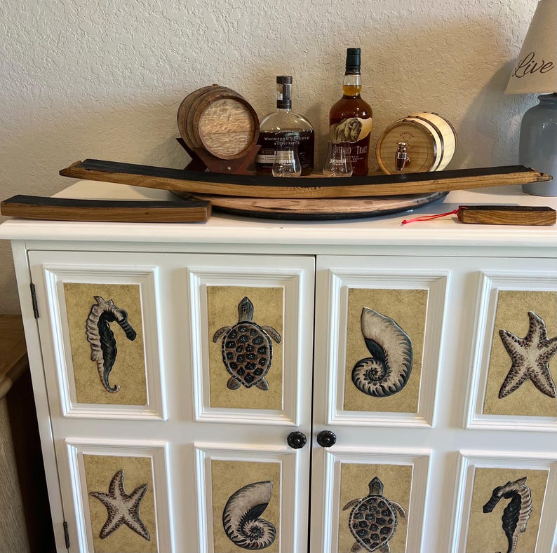 Puede incluir: Un mueble blanco con obras de arte de tem&aacute;tica marina en las puertas, incluyendo caballitos de mar, tortugas y estrellas de mar. Encima, un barril de madera, botellas de licor y una pieza decorativa de madera crean una est&eacute;tica r&uacute;stica.