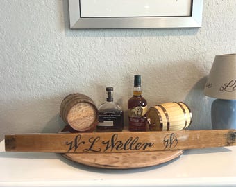 May include: A wooden sign with the text "W L Weller" in black script, along with two small wooden barrels and two bottles of liquor. The sign is on a wooden tray.