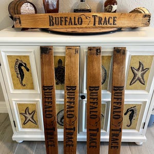 May include: Wooden sign and staves with "BUFFALO TRACE" text. The sign is made from a barrel stave and the staves are vertical. The sign and staves are brown with black lettering. The background includes liquor bottles and a white cabinet.