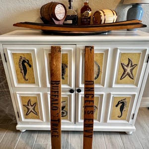 May include: A white cabinet with a decorative wooden barrel stave handle. The cabinet doors feature framed sea creature art. A wooden tray holds small barrels and bottles of liquor. The staves read "BUFFALO TRACE" and "ANGEL'S ENVY".