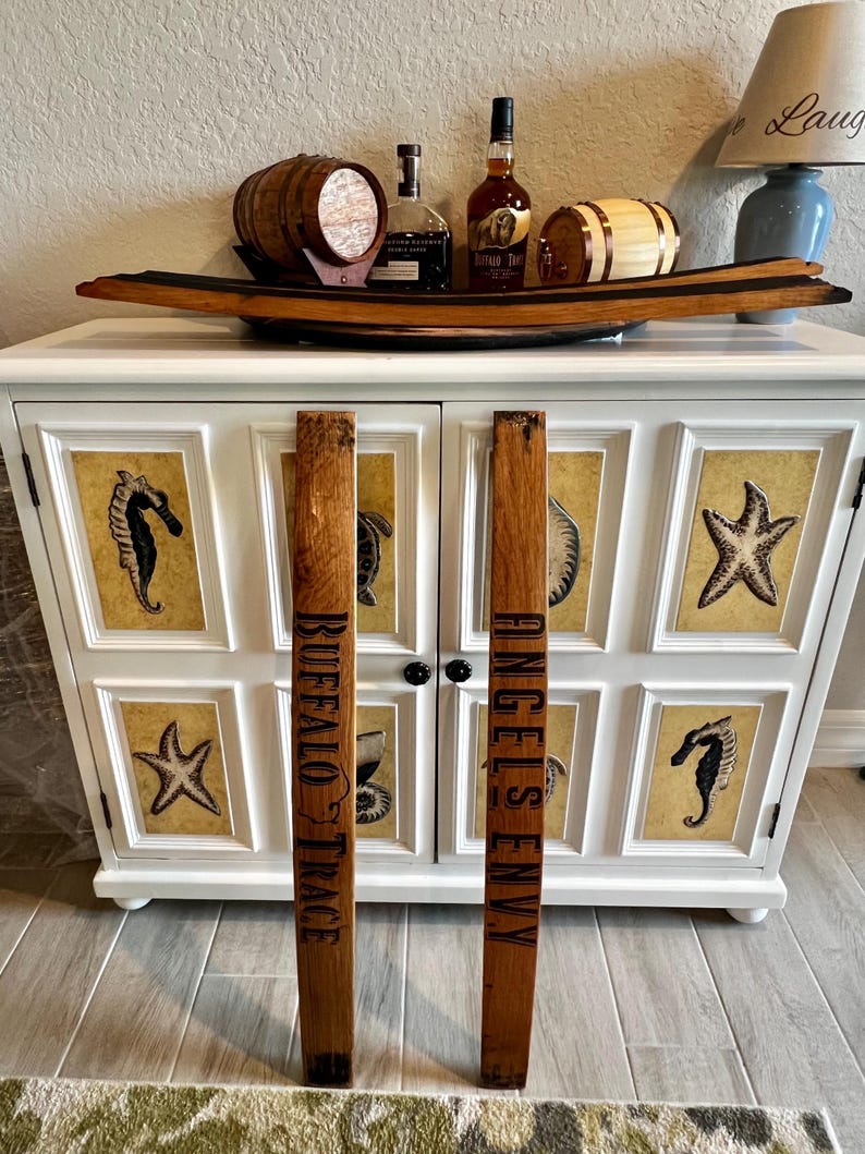 May include: A white cabinet with a decorative wooden barrel stave handle. The cabinet doors feature framed sea creature art. A wooden tray holds small barrels and bottles of liquor. The staves read "BUFFALO TRACE" and "ANGEL'S ENVY".