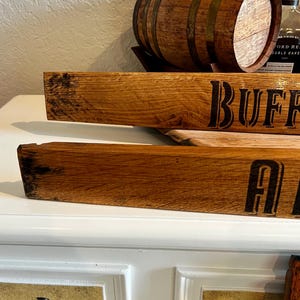 May include: Wooden barrel stave with the word "BUFF" in black lettering. The stave is made of light brown wood with a textured surface. A small wooden barrel with metal bands sits above the stave. The image suggests a rustic, vintage theme.