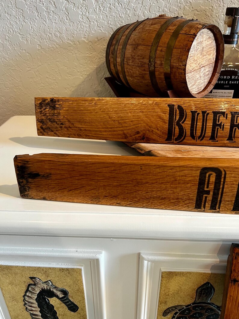 May include: Wooden barrel stave with the word "BUFF" in black lettering. The stave is made of light brown wood with a textured surface. A small wooden barrel with metal bands sits above the stave. The image suggests a rustic, vintage theme.