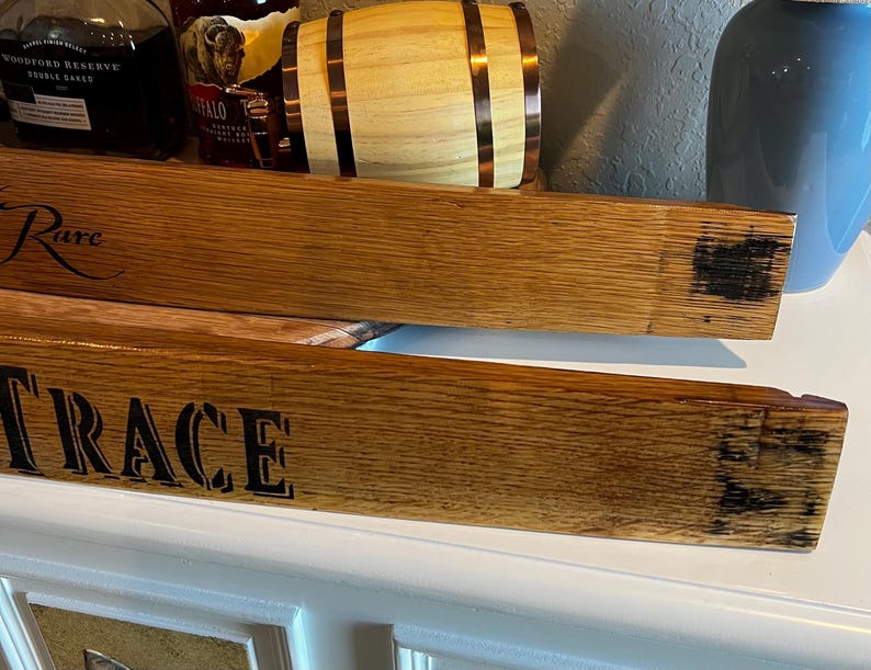 Op de afbeelding: Twee houten duigen met de woorden "Rare" en "Trace" in zwart ge&euml;tst. De duigen zijn lichtbruin van kleur met donkere verkoolde uiteinden. Een klein houten vat en flessen drank staan op de achtergrond.