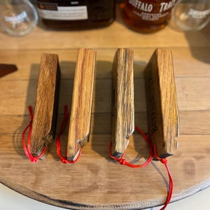 May include: Four wooden whiskey stave coasters with red string attached. The coasters are made from reclaimed wood and have a natural finish. The Buffalo Trace logo is visible on one coaster. The coasters are arranged on a wooden surface.