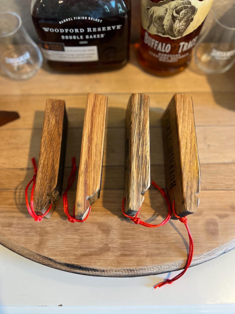 May include: Four wooden whiskey stave coasters with red string attached. The coasters are made from reclaimed wood and have a natural finish. The Buffalo Trace logo is visible on one coaster. The coasters are arranged on a wooden surface.