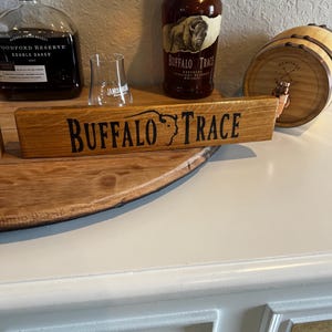 Puede incluir: Un letrero de madera con las palabras "BUFFALO TRACE" en letras negras. El letrero se coloca frente a una botella de bourbon Buffalo Trace y un vaso peque&ntilde;o. Tambi&eacute;n es visible un peque&ntilde;o barril de madera.