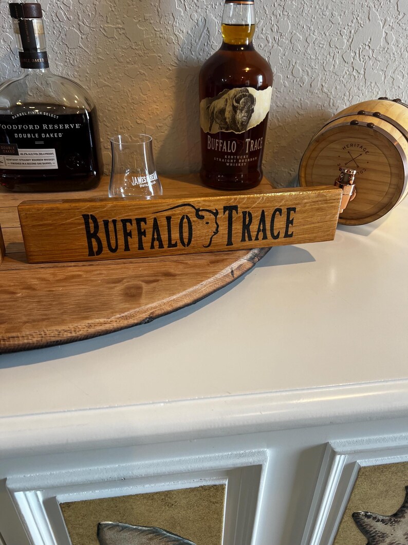 Puede incluir: Un letrero de madera con las palabras "BUFFALO TRACE" en letras negras. El letrero se coloca frente a una botella de bourbon Buffalo Trace y un vaso peque&ntilde;o. Tambi&eacute;n es visible un peque&ntilde;o barril de madera.