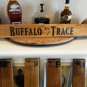 May include: A wooden sign with the words "BUFFALO TRACE" in black lettering, with three bottles of bourbon and two glasses in the background. The sign is made of light brown wood and has a curved shape.