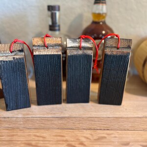 May include: Four rectangular wooden barrel stave ornaments with a dark charred front and a natural wood top. Each ornament has a red string for hanging. The background includes whiskey bottles and wooden barrels.