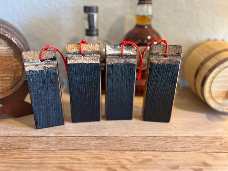 May include: Four rectangular wooden barrel stave ornaments with a dark charred front and a natural wood top. Each ornament has a red string for hanging. The background includes whiskey bottles and wooden barrels.