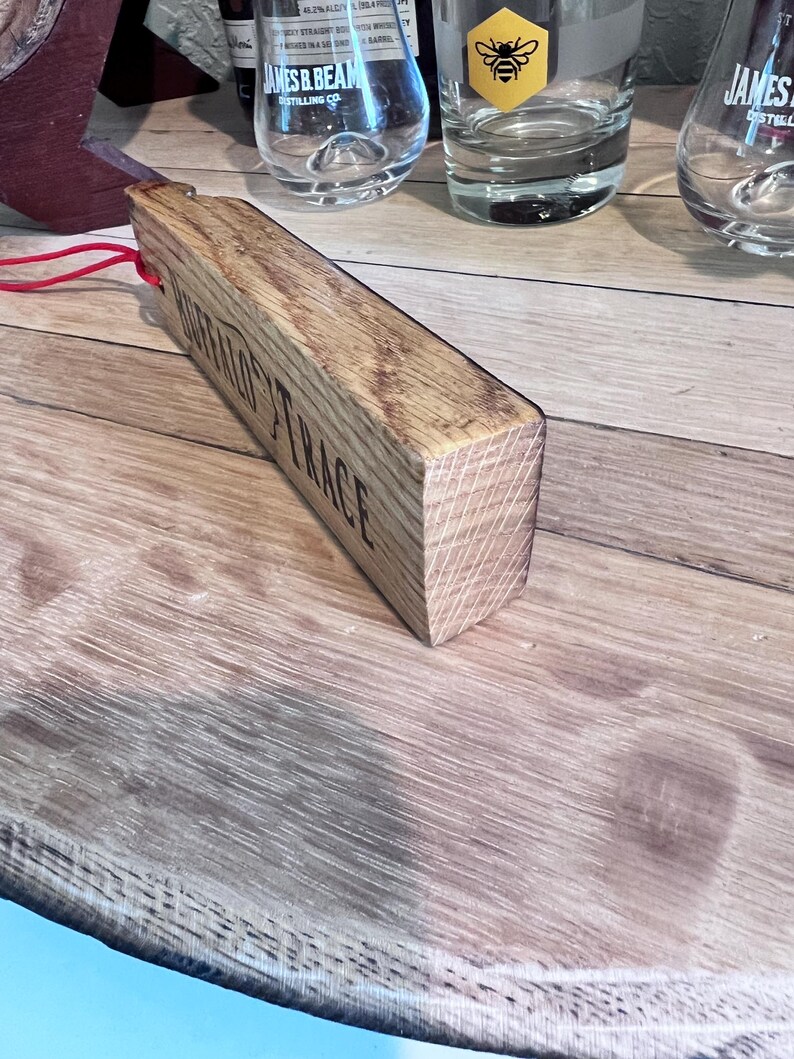 May include: A wooden bottle stopper with the text "BUFFALO TRACE" engraved on the side. The stopper is rectangular and made of light brown wood. A red string is attached to the stopper. Several glasses are in the background.