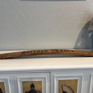 May include: A curved, wooden stave with the words "BUFFALO TRACE" burned into the surface. The stave is brown and appears to be from a barrel. The background includes a blue lamp and a white cabinet.
