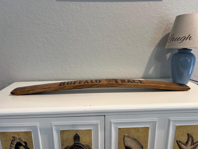May include: A curved, wooden stave with the words "BUFFALO TRACE" burned into the surface. The stave is brown and appears to be from a barrel. The background includes a blue lamp and a white cabinet.