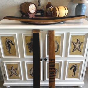 Puede incluir: Un mueble blanco con arte enmarcado con tem&aacute;tica marina. Encima, una duela de barril de madera sostiene barriles decorativos y botellas de licor. Una duela dice "Fireball Whiskey."