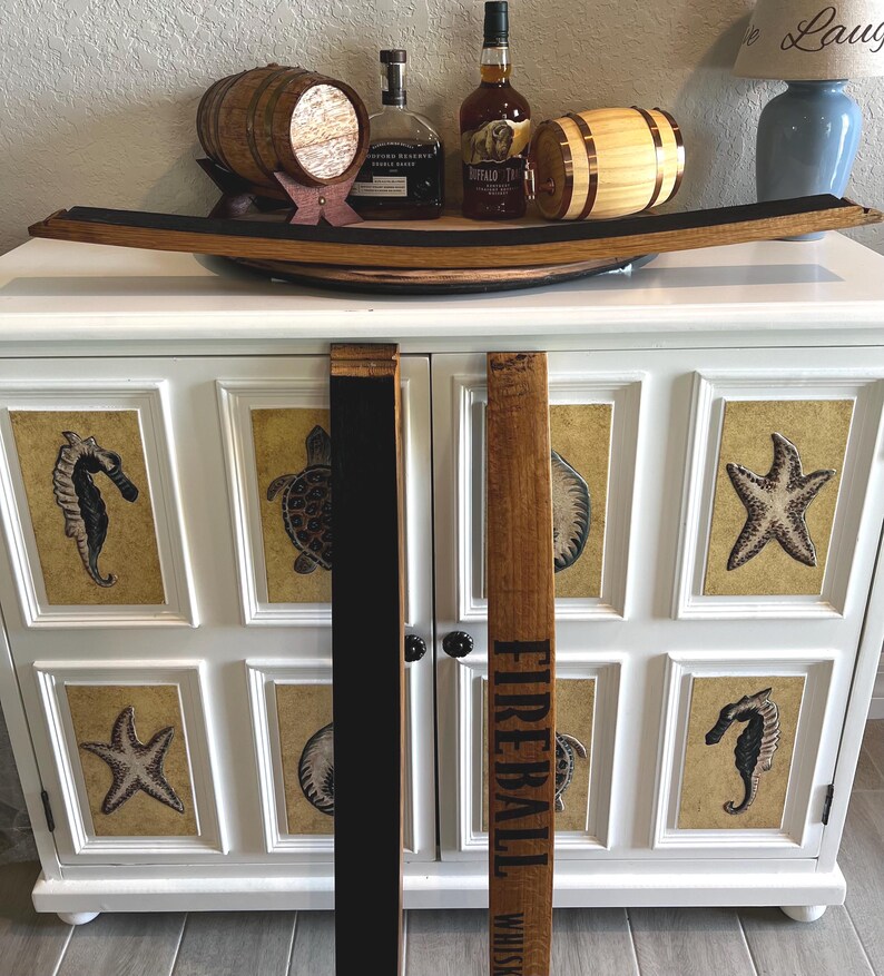 Puede incluir: Un mueble blanco con arte enmarcado con tem&aacute;tica marina. Encima, una duela de barril de madera sostiene barriles decorativos y botellas de licor. Una duela dice "Fireball Whiskey."