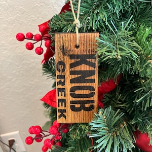 May include: A wooden Christmas tree ornament with the words "KNOB CREEK" printed in black. The ornament is rectangular and hangs from a beige string. The ornament is surrounded by green pine needles, red berries, and red decorative leaves.