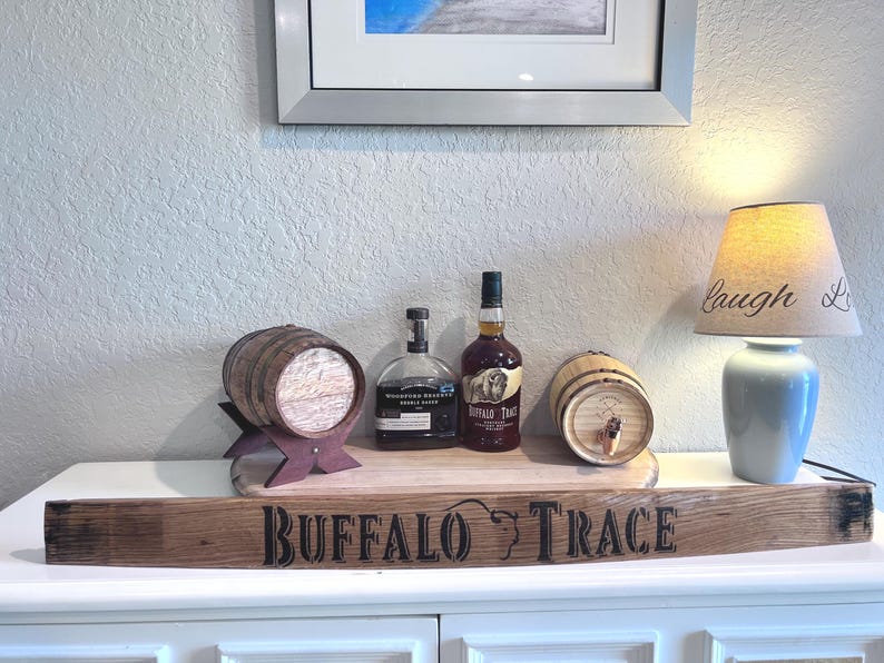 May include: A display featuring a wooden barrel, bottles of liquor, and a lamp with a lampshade that says "Laugh Love". The display sits on a wooden plank with the words "Buffalo Trace" on it.