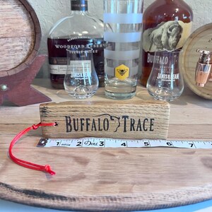 May include: A wooden block with "BUFFALO TRACE" text, a red string, and a measuring tape. Clear glasses, a bottle of bourbon, and a small barrel are in the background. The tape measure reads 6 inches.