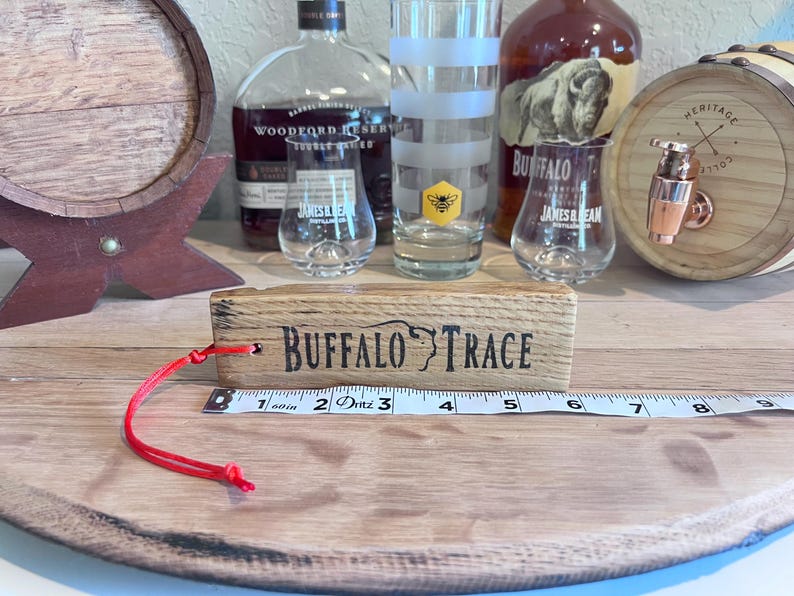 May include: A wooden block with "BUFFALO TRACE" text, a red string, and a measuring tape. Clear glasses, a bottle of bourbon, and a small barrel are in the background. The tape measure reads 6 inches.