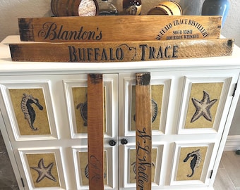 May include: Wooden signs with the text "Blanton's" and "Buffalo Trace" are displayed on a white cabinet. The cabinet doors feature sea-themed artwork. Two wooden planks with initials are in front of the cabinet. Bottles and a small barrel are on top.