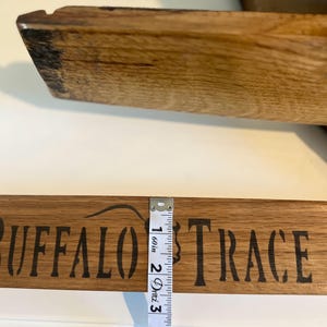 Puede incluir: Una secci&oacute;n de tabla de madera con las palabras "BUFFALO TRACE" estampadas en negro. Una cinta m&eacute;trica muestra que la tabla tiene aproximadamente 2,5 cm de grosor. La madera tiene un aspecto natural y envejecido.