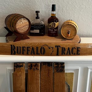 May include: A wooden sign with the text "BUFFALO TRACE" and a buffalo head logo. The sign is displayed on a white cabinet, with two small wooden barrels and two bottles of liquor on top. The barrels are made of wood and have a tap.